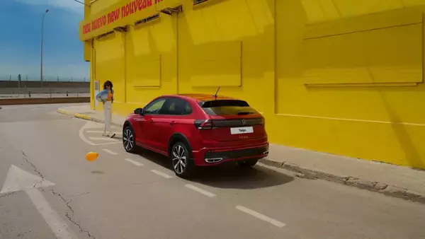 VW Taigo rouge sur le bord de la route devant un bâtiment jaune, vue arrière et latérale, une femme marche vers le vhicule.